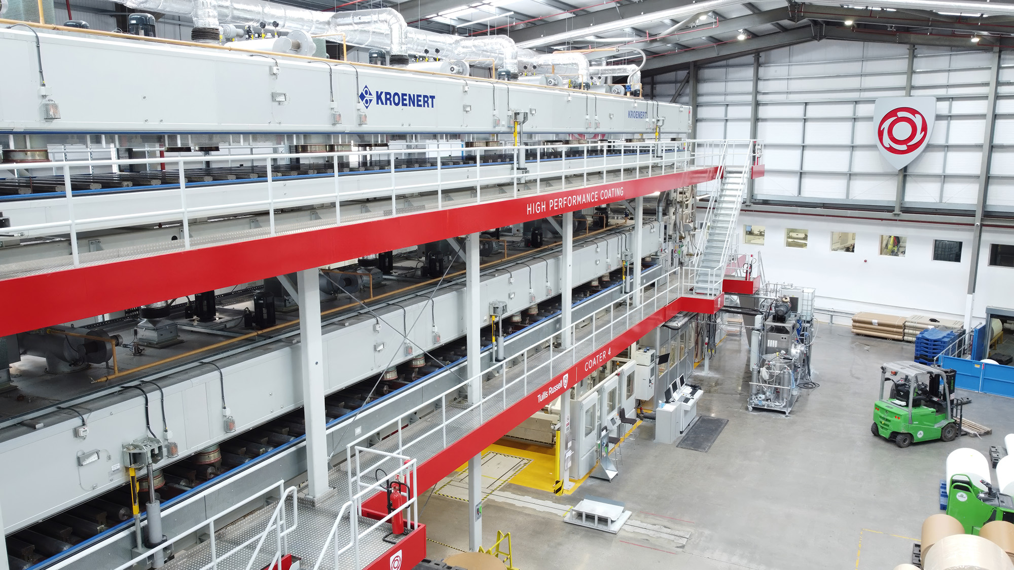 A large three-story grey and red industrial machine stands in a factory hall, viewed from an elevated left corner. Below, green forklifts move large paper reels across the floor.